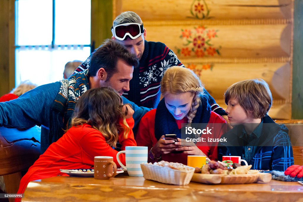 Friends having lunch after skiing, using a smart phone Group of friends wearing winter clothes sitting in a mountain restaurant after skiing, having lunch, woman using a smart phone. Winter Stock Photo