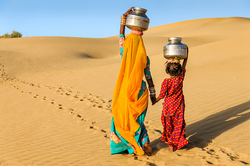 Indian mother and her little daughter crossing sand dunes and carrying on their heads water from local well, Thar Desert, Rajasthan, India. Rajasthani women and children often walk long distances through the desert to bring back jugs of water that they carry on their heads. 