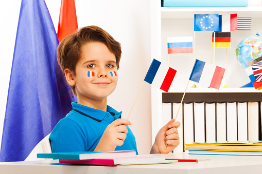 Smiling boy sitting at desk in the classroom holding two French flags in his hands