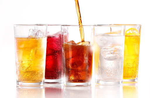 A row of five soft fizzy drinks, with one still being poured.  Drinks are shot with ice and are shot on white reflective background