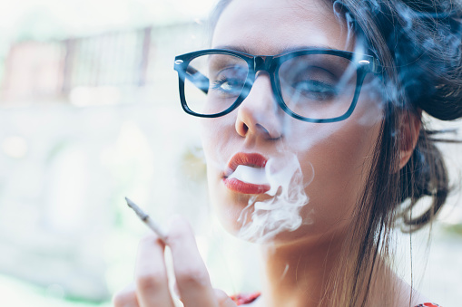 Beautiful young woman with eyeglasses and red lips smoking cigarette. Looking at camera. Close up.