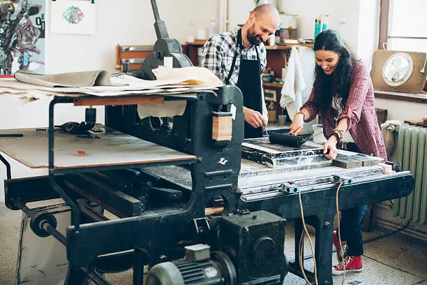 Man and woman lithography workers handmaking new products at their workshop. Using printig press and tools in printing house. Man holding a brush, woman using a roller.