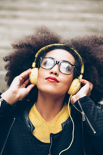 Young attractive girl listening music with yellow headphones, looking up. Wear yellow pullover, black jacket and blue jeans, and black eyeglasses. Hands on the headphones.
