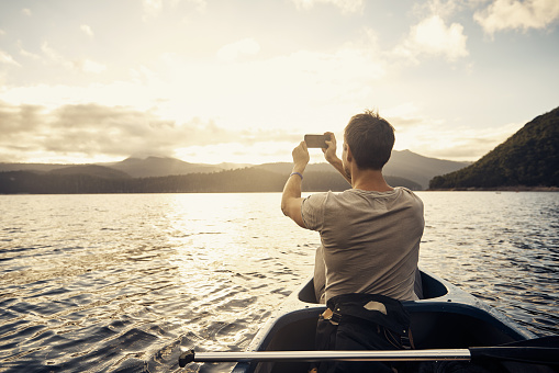 Rearview shot of a man taking a picture with his phone while out for a boat ride on the lake