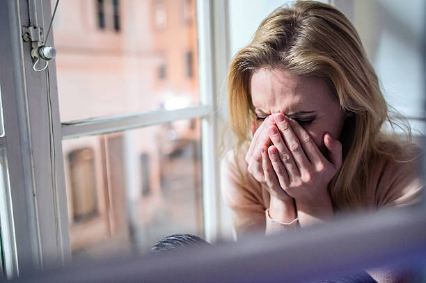 Woman sitting on windowsill, looking out of window, crying Beautiful blond woman sitting on window sill at night, looking out of window, crying women crying stock pictures, royalty-free photos & images