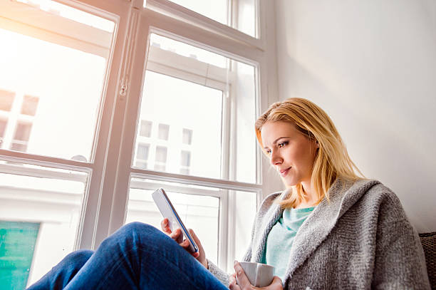 istock Woman sitting on window sill with smartphone and coffee 521086820