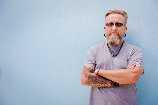 Mature rock star with tatoo and piercing posing, isolated on blue wall background