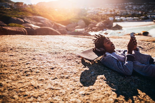 African American skater lying down, listening to music from his smartphone with headphones and resting his head on his skateboard on the rocks of a seaside during sunset