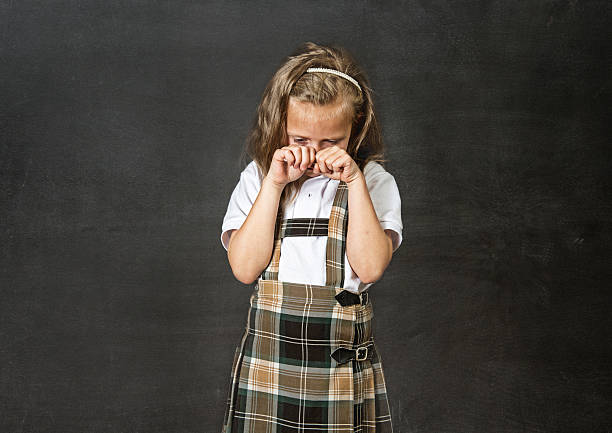 junior schoolgirl crying in children education stress and bullying victim young sweet junior schoolgirl with blonde hair crying sad standing isolated in front of classroom blackboard holding chalk in her little hand wearing school uniform in children education stress embarrassment skirt photos stock pictures, royalty-free photos & images