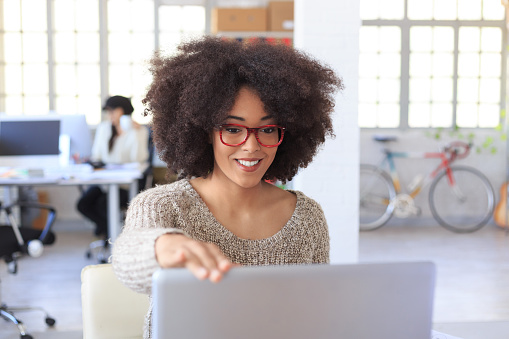 Smiling young woman with red eyeglasses finishing her work at the office and closing laptop. Looking at it. As background desk with computers and colleagues, bike, guitar and wall with windows.