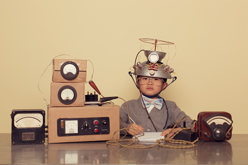 A young Japanese boy sits at office desk searching for successful ideas by using a mind reading helmet. He is working on ideas in the inner most parts of the brain. He is wearing a cardigan and bow tie.