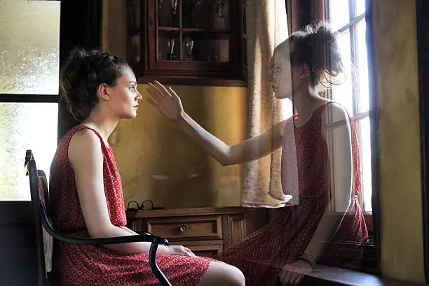 Pensive young woman looking throuth the window. Wears red dress and sitting on chiar in vintage house. Her ghost in front of her trying to touch her.