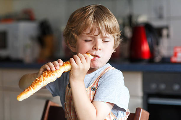 Close-up of a young boy in retro cap holding a loaf of bread near a haystack. Rustic lifestyle in countryside, blue sky background.