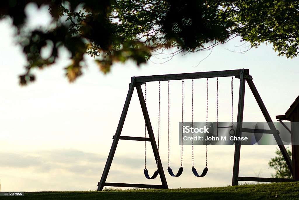 Swingset at sunset, silhouette A wooden swings is backlit by a late evening sky framed by leaves in the foreground. Back Lit Stock Photo