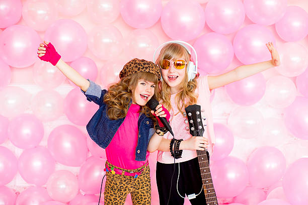 two little girl singing a song with a microphone and a guitar on a background of pink balloons