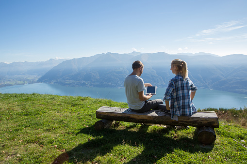 Young couple sitting on a bench surrounding a beautiful mountain/lake landscape using a digital tablet.