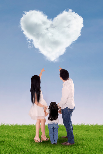 Rear view of three member happy family looking at cloud shaped heart in field
