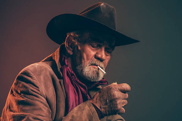 istock Senior cowboy with gray beard and brown hat lighting cigarette. 521247351