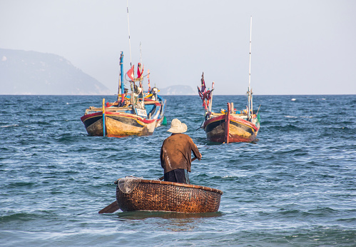 Doc Let, Vietnam - February 12, 2016: A fisherman at Doc Let near Nha Trang, Vietnam is driving his boat through the waves. The boat is round and it is woven from bamboo. In the background are fisherboats.