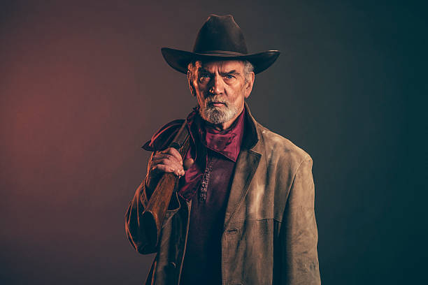 Senior cowboy with gray beard and brown hat holding rifle. Old rough western cowboy with gray beard and brown hat holding rifle. Low key studio shot. old guns stock pictures, royalty-free photos & images
