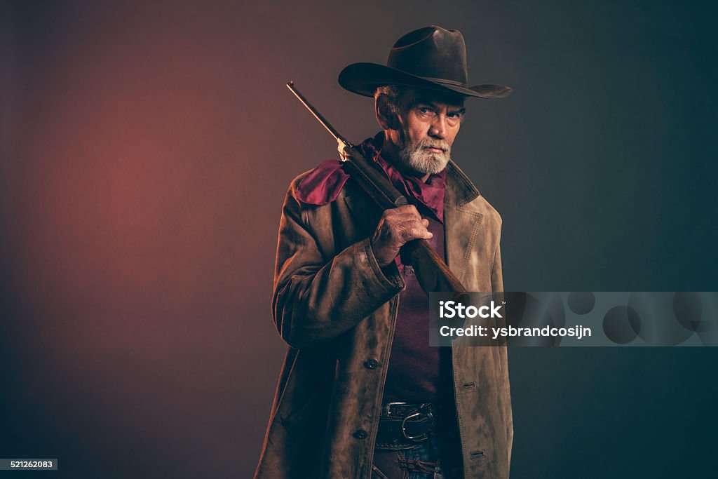 Senior cowboy with gray beard and brown hat holding rifle. Old rough western cowboy with gray beard and brown hat holding rifle. Low key studio shot. Cowboy Stock Photo