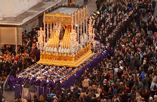 Malaga, Spain - March 22, 2016: eight rows of men dressed in purple robes, carry a heavy float with a statue of Virgin Mary, richly decoarted with golden ornaments and burning candles, in the Easter procession of the Rescate brotherhood on holy Tuesday. The procession is watched by many spectators . 
