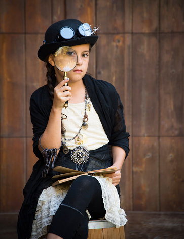 Young girl dressed in SteamPunk style holds a large magnifying glass looking back at the camera