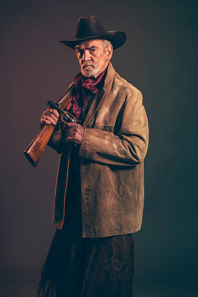 Old rough western cowboy with gray beard and brown hat holding rifle and revolver. Low key studio shot.
