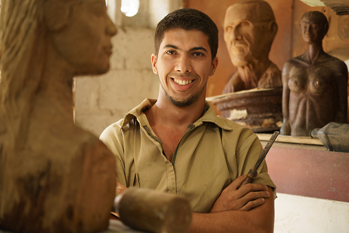 Portrait of man working, young student in art class, learning crafts profession, working with wooden statue and looking at camera