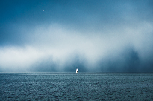 A sailboat sits on the horizon of the ocean under misty clouds.