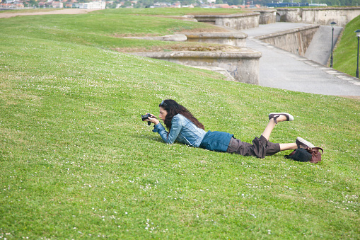 brunette adult tourist woman with blue jeans jacket and brown trousers lying on the green grass with camera travel photo, as photographer taking picture of flower in Gijon park Asturias Spain