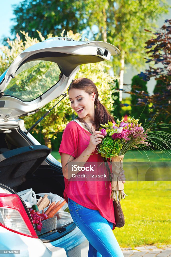 Young woman standing outdoor by car, holding flowers Cheerful young woman came back from shopping, standing outdoor by the car trunk full of shopping bags and holding bouquet of flowers. Car Stock Photo