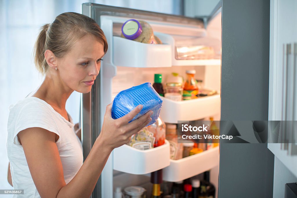 Pretty, young woman in her kitchen by the fridge Is this still fine? Pretty, young woman in her kitchen by the fridge, looking at the expiry date of a product she took from her fridge - Obsolete Stock Photo