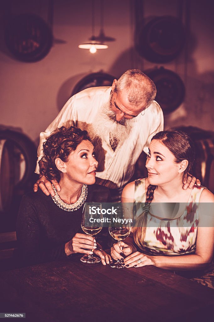 Friends in Wine Cellar Senior winemaker with white long beard standing behind two adult female friends who sitting at the table in winery cellar, tasting white wine and talking to him. Vertical color image. Goriška Brda, Slovenia, Europe. Active Seniors Stock Photo