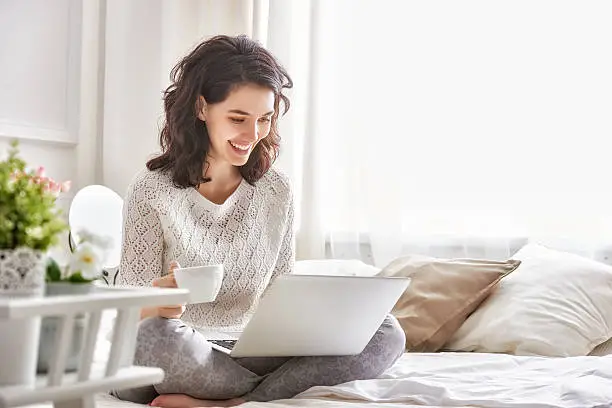 Happy casual beautiful woman working on a laptop sitting on the bed in the house.