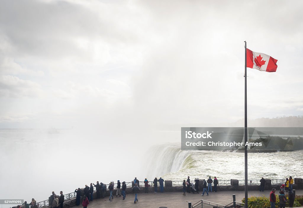 Niagara Falls With Tourists and Canadian Flag Under the Maple Leaf Flag, tourists line the Niagara river bank on the Canadian side of the famous Horseshoe Falls to admire the view. Niagara Falls Stock Photo