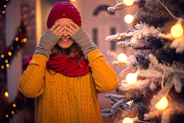 Happy caucasian woman outdoors in front of house. Standing by christmas tree and covering eyes with her hands waiting for surprise. Wearing knitted sweater, hat and scarf. House, yard and tree are decorated with festive string lights. Evening or night with beautiful yellow lights lightning the scenes.
