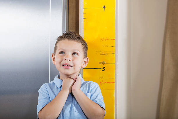 Little boy measuring his height on growth chart An Hispanic 5 year old boy standing next to a height chart. He is looking away, with a candid expression, hoping he has gotten taller. boy height measure stock pictures, royalty-free photos & images