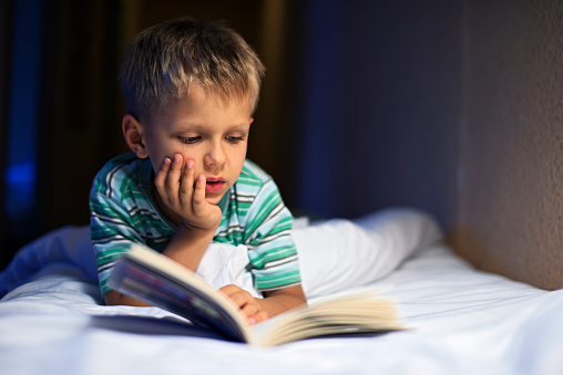 Little boy aged 6 is reading book in bed before going to sleep. The book is very interesting and the boy is quite lost in it.