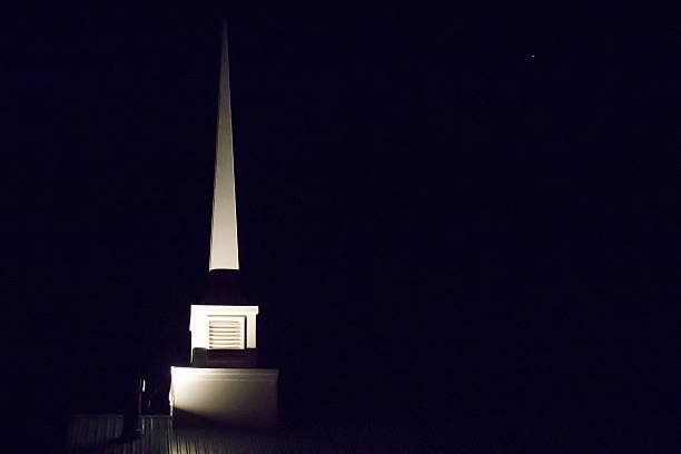 Image of a triangular shaped church steeple in the darkness of night.  The place of worship is white and is illuminated by a light below.  The sky is dark black.