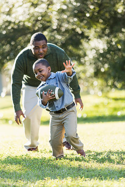 African American father and son playing football An African American man in his 40s playing football with his 5 year old son. It is a bright, sunny spring day. They are outdoors in the park or the yard, trees out of focus in the background. The boy is holding the ball and his dad is standing behind him. chasing photos stock pictures, royalty-free photos & images