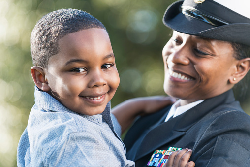 An African American woman wearing a naval officer uniform holding her 5 year old son. The focus is on the little boy who is smiling and looking at the camera.