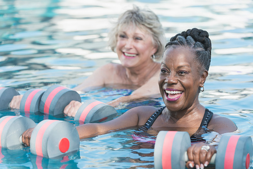 Two senior woman, one Caucasian and the other African American, standing in a swimming pool holding dumbbells, taking an exercise class doing water aerobics. They are smiling, having a good time while they stay in shape. Focus is on the black woman in the foreground.