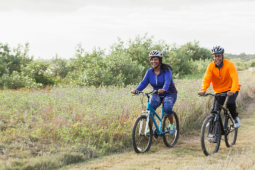 A mature, African American couple riding mountain bikes in a state park. They are wearing helmets, cycling side by side, smiling.