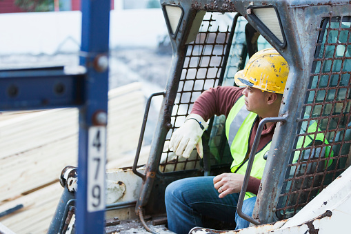 High angle view of an Hispanic construction worker at a job site, sitting in a backhoe, wearing a yellow hardhat and safety vest.