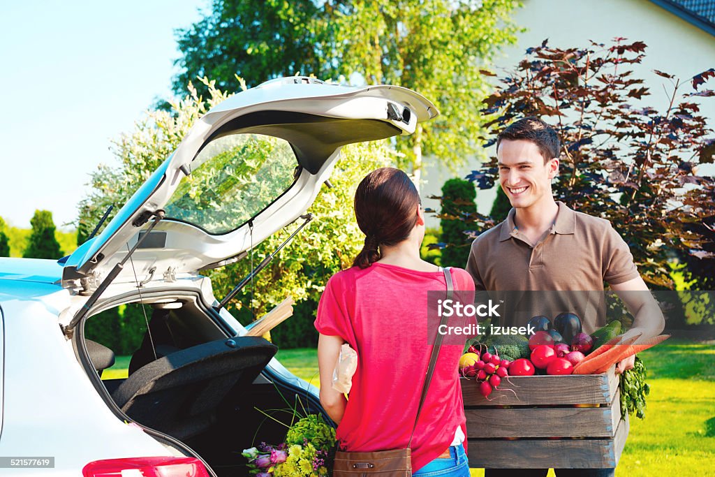 Young couple standing outdoor by car, came back from shopping Cheerful young couple came back home from shopping, talking outdoor by the car trunk, the man holding a box of vegetables.  Adult Stock Photo