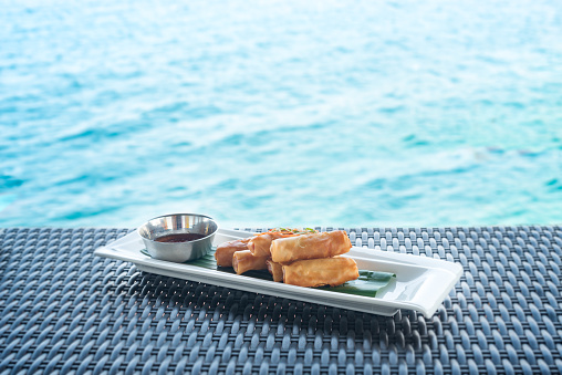 Vegetable fried spring rolls served with bittersweet sauce on wicker table and water background