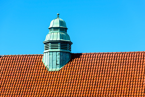 Small copper tower on top of a tiled roof with blue sky in background.