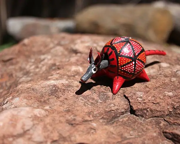 Mexican bobble head toy red armadillo on a rock.  The red wooden toy is painted with a geometric pattern, with dots, lines and triangles.  These toys are commonly sold in Mexico.  This close up shot of the bobble head has it sitting on a pink rock.  