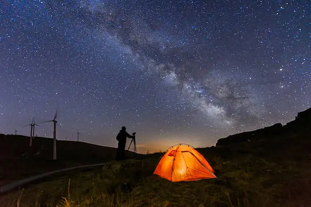 A Photographer is standing on the top of the hill next to the Milky Way galaxy with his hands on the tripod.
